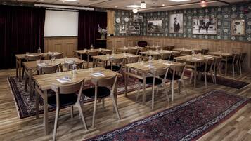 A conference room with wooden tables and chairs, a projector screen, and a stage with curtains.