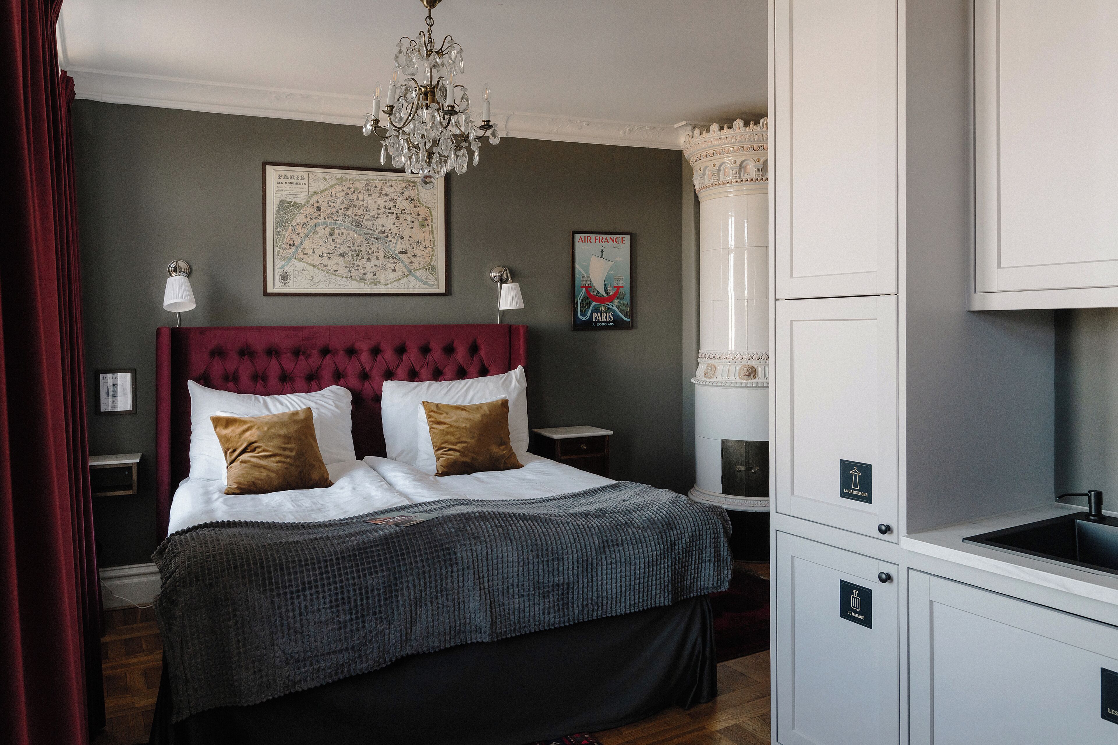 Un dormitorio con cabecera capitonada, cama con almohadas blancas y marrones, un mapa y un cartel en la pared, una araña y una zona de cocina integrada.