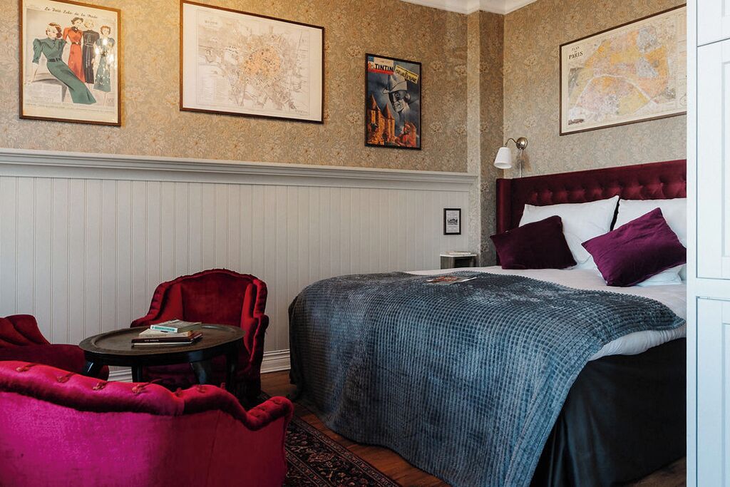 Un dormitorio con una cama, un sillón rojo, una mesita y tres cuadros enmarcados en la pared.
