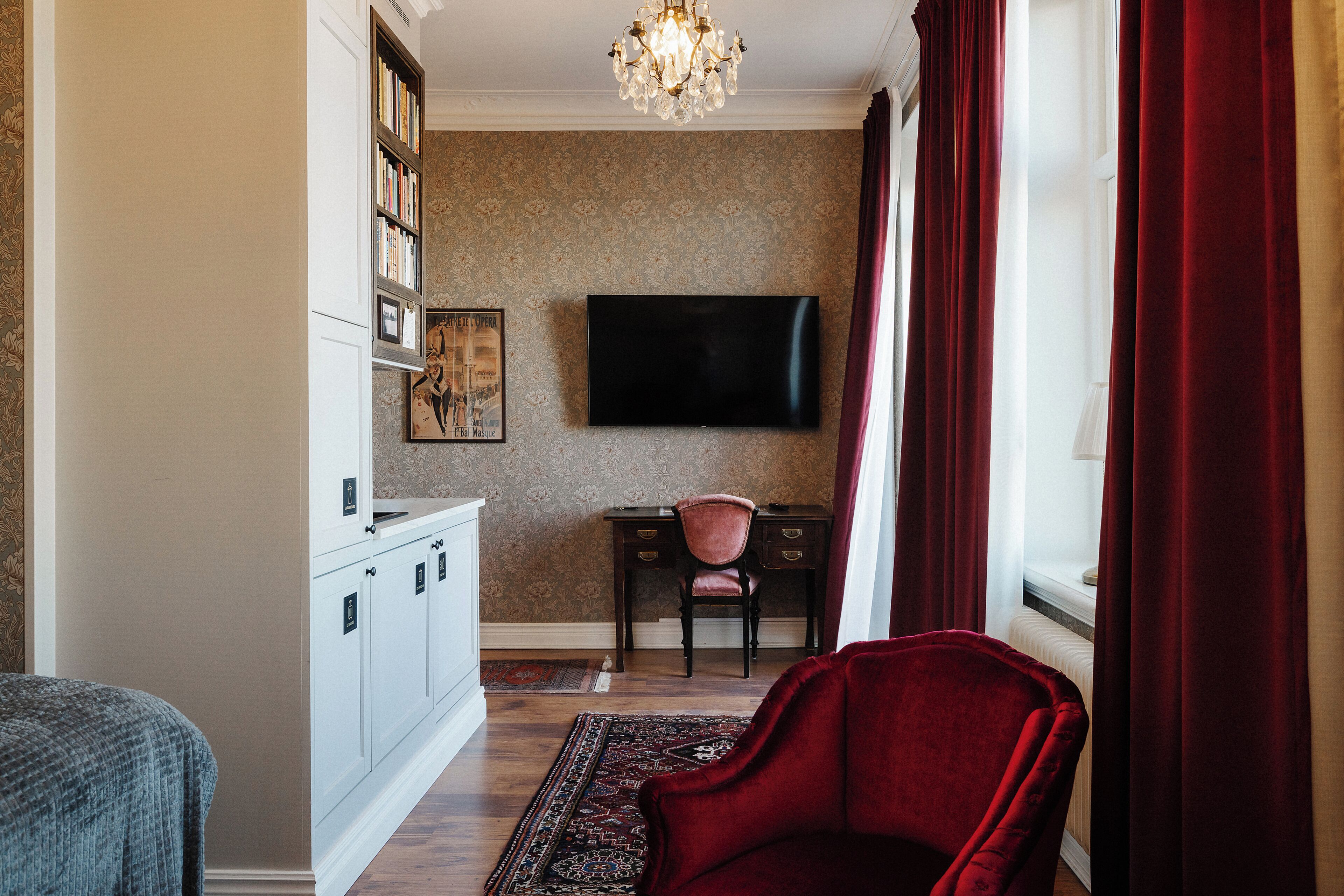 Una habitación con televisión montada en la pared, un escritorio con silla, una silla roja y una alfombra estampada.