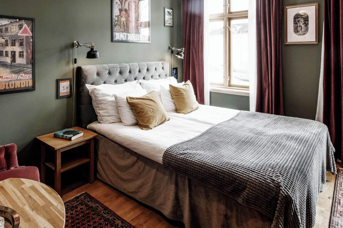 Ein ordentlich eingerichtetes Schlafzimmer mit einer gesteppten Kopfteil, einem großen Bett mit vielen Kissen, einem Nachttisch mit einem Buch und einem Fenster mit Vorhängen.