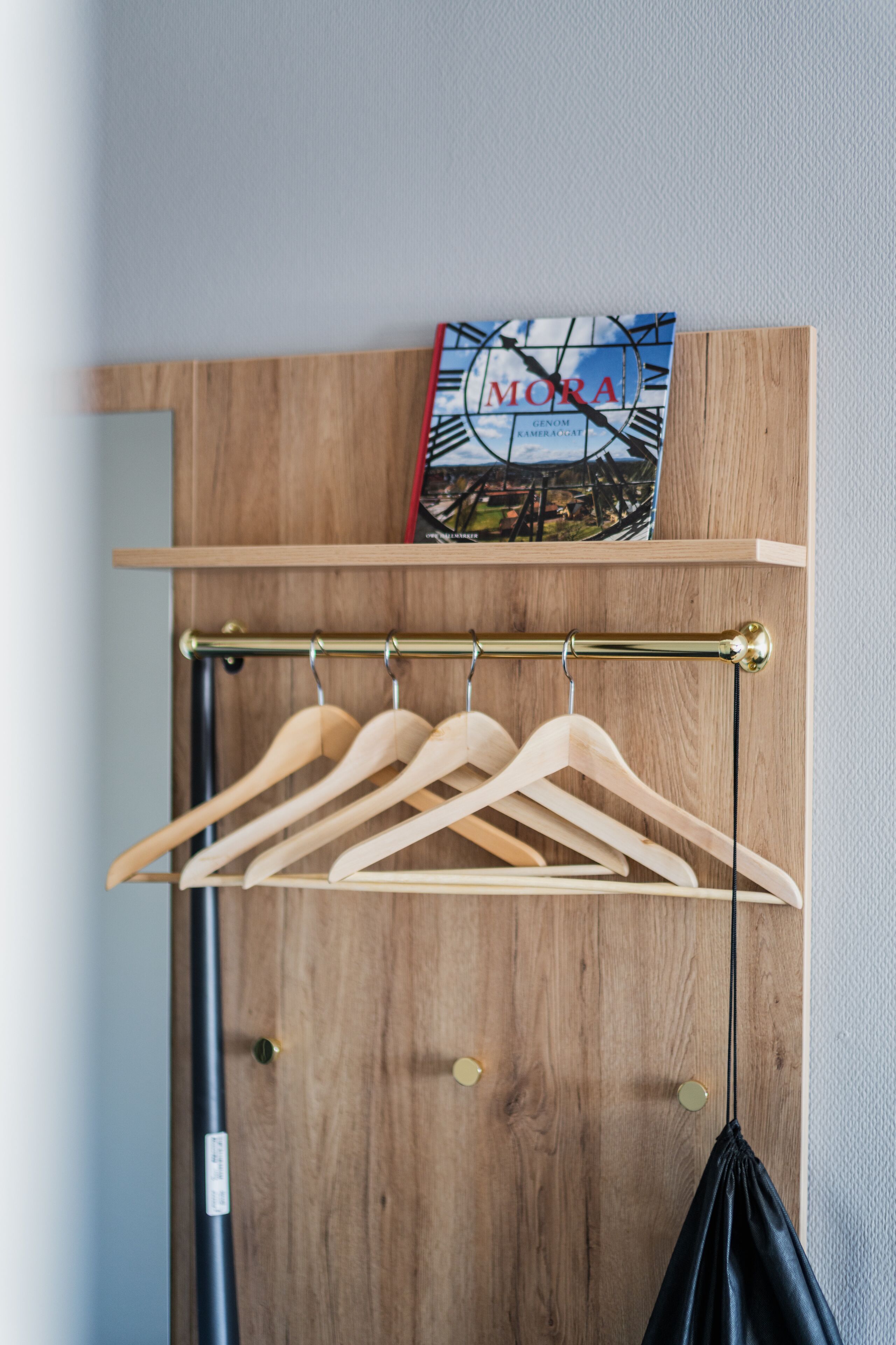 A wooden wardrobe with a hanging rack above it, a book titled "MORA" on top, and a black bag hanging below.