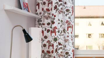 Quarto com escrivaninha, luminária, cadeira, janela com vista para edifícios e cortina estampada com motivos florais.