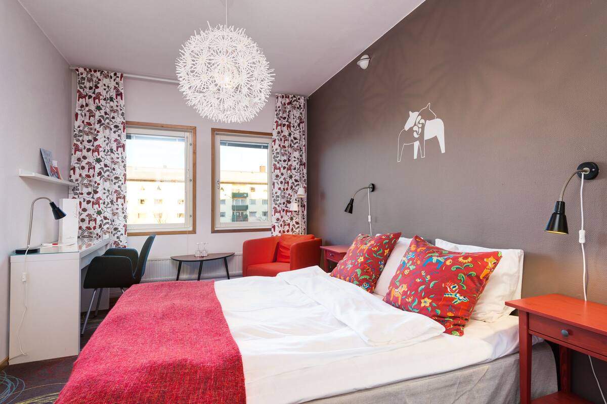 A hotel room with a bed, a desk, a chair, a red armchair, a small table, and a decorative wall piece.