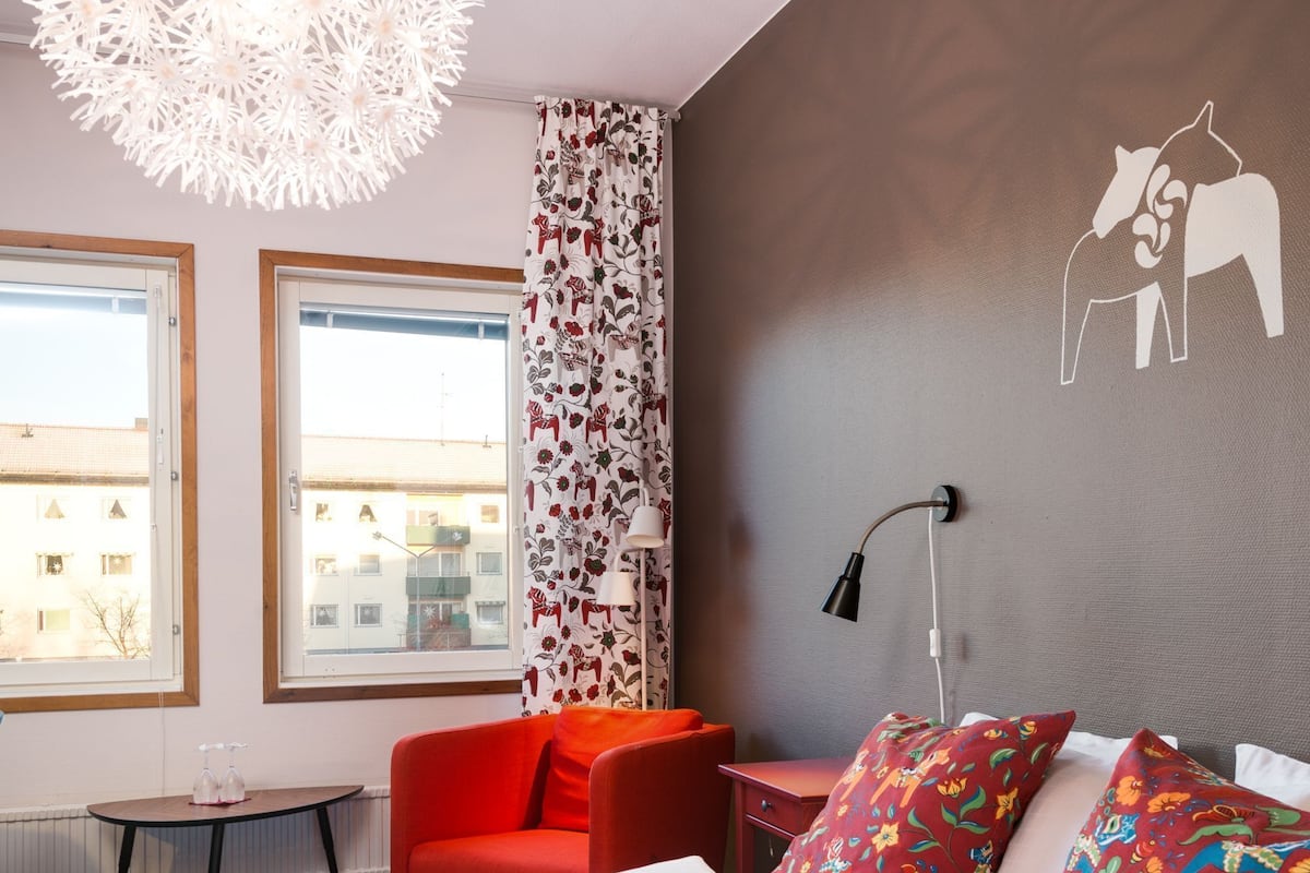 A hotel room with a large bed, a red chair, a small table, a hanging light fixture, and a wall with a dog silhouette.