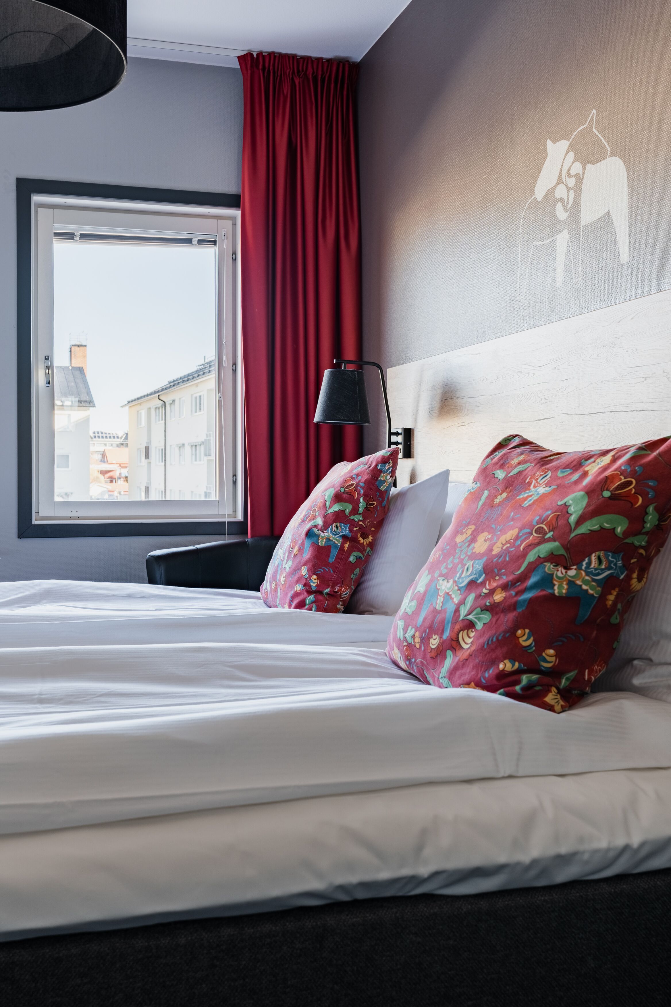 A hotel room with a bed, red curtains, a city view, and a wall art of a horse.