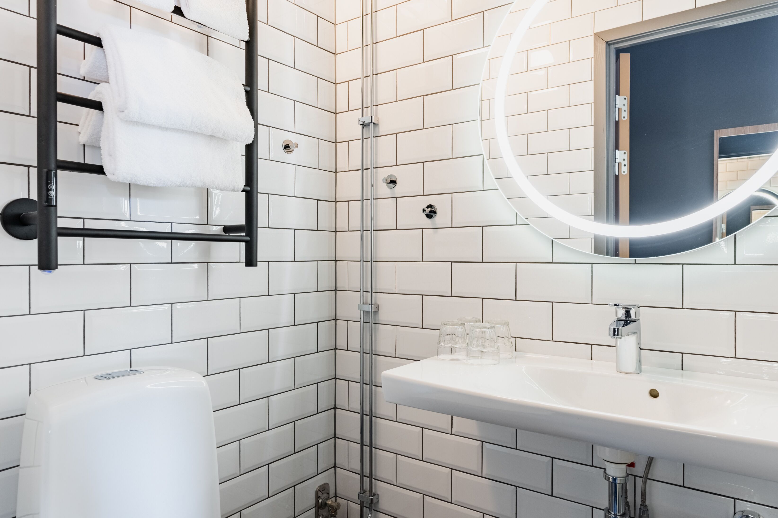 Ein modernes Badezimmer mit weißen Fliesen, einer Duschkabine aus Glas, einem schwarzen Handtuchhalter, einem Waschbecken mit Spiegel und einer Seifenspender.