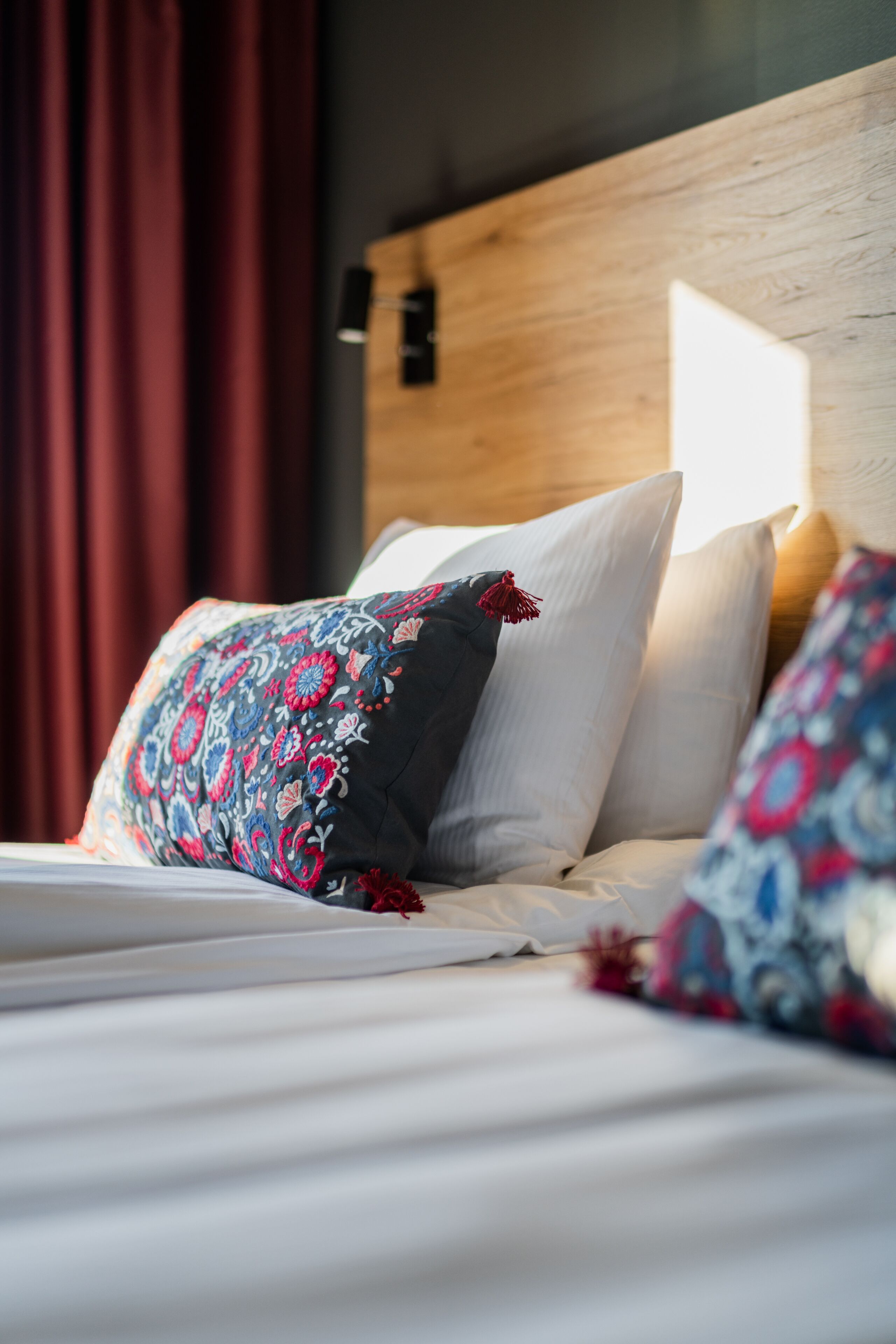 A bed with white linens and patterned pillows, a wooden headboard, and a red curtain in the background.