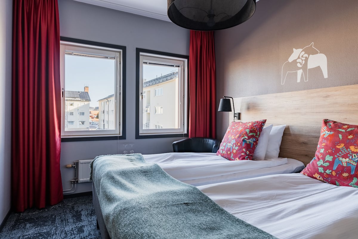A hotel room with a bed, two windows, a radiator, a lamp, and a wall with a horse and rider mural.