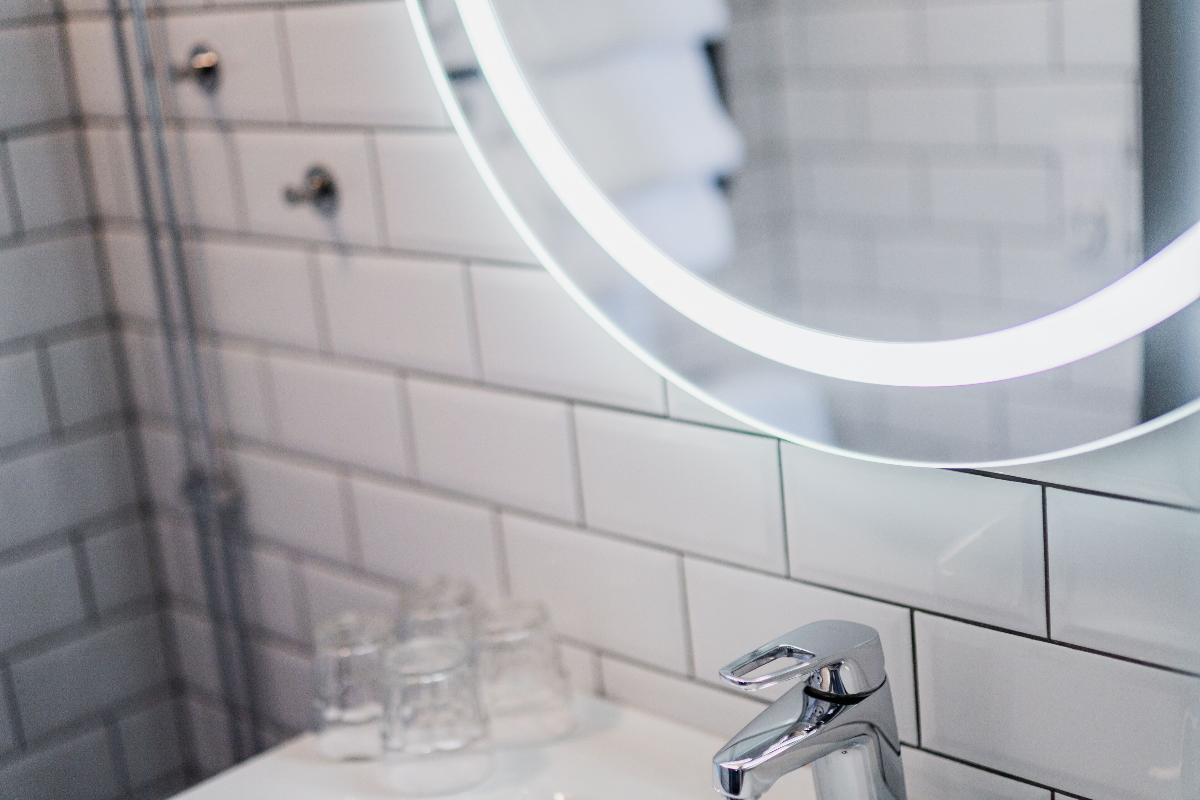 Une salle de bain moderne avec un grand miroir rond, un robinet chromé et des murs recouverts de carreaux blancs.