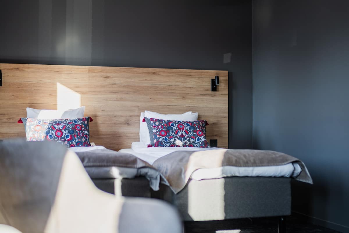 A modern hotel room with a wooden headboard, two beds with patterned pillows, and a grey armchair.