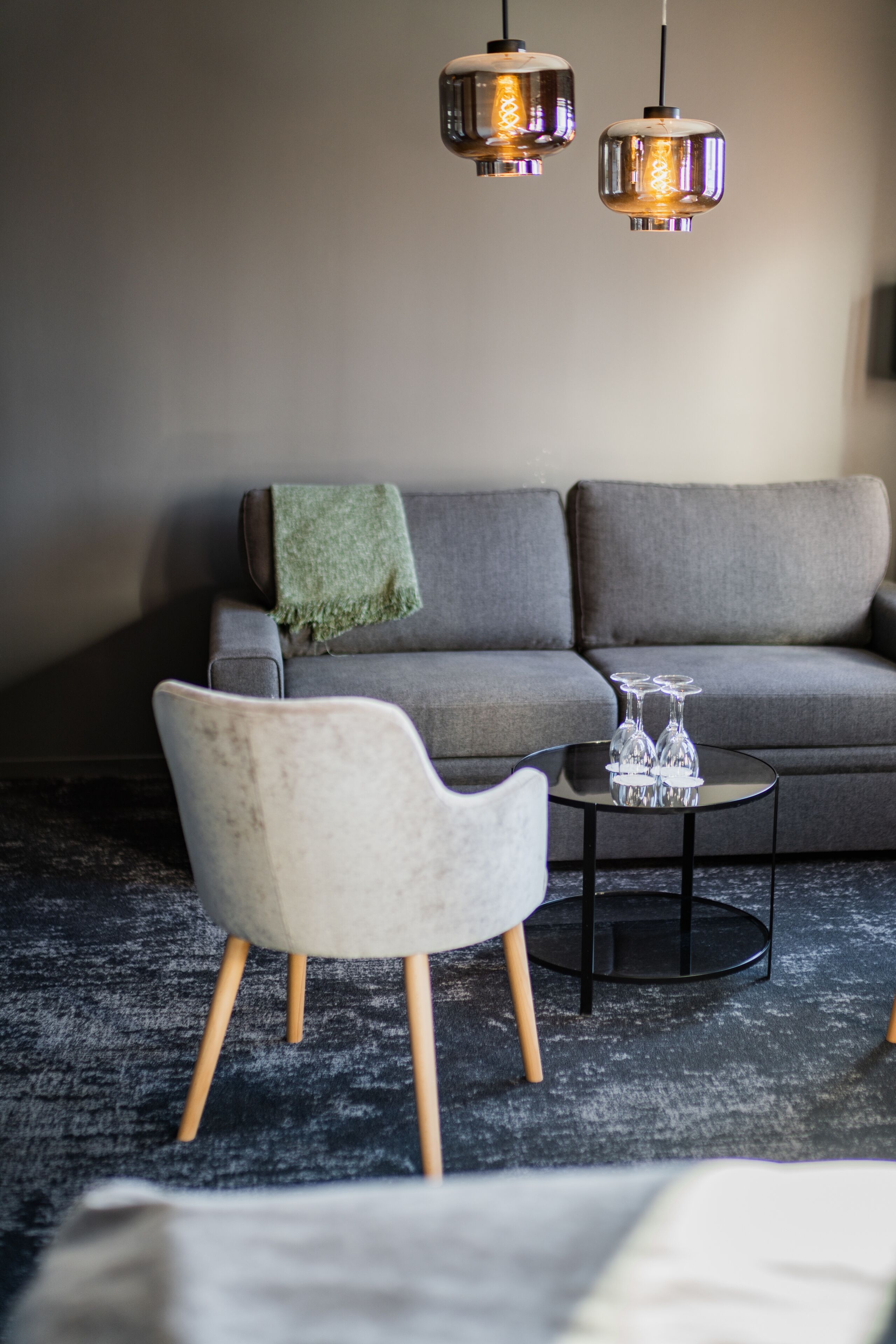 A modern living room with a grey sofa, a white armchair, a round coffee table with wine glasses, and two hanging pendant lights.