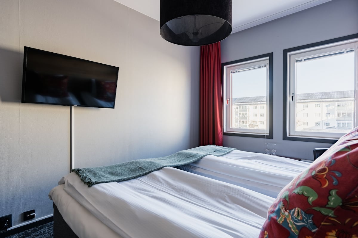 A hotel room with a bed, a TV mounted on the wall, and two windows with curtains.