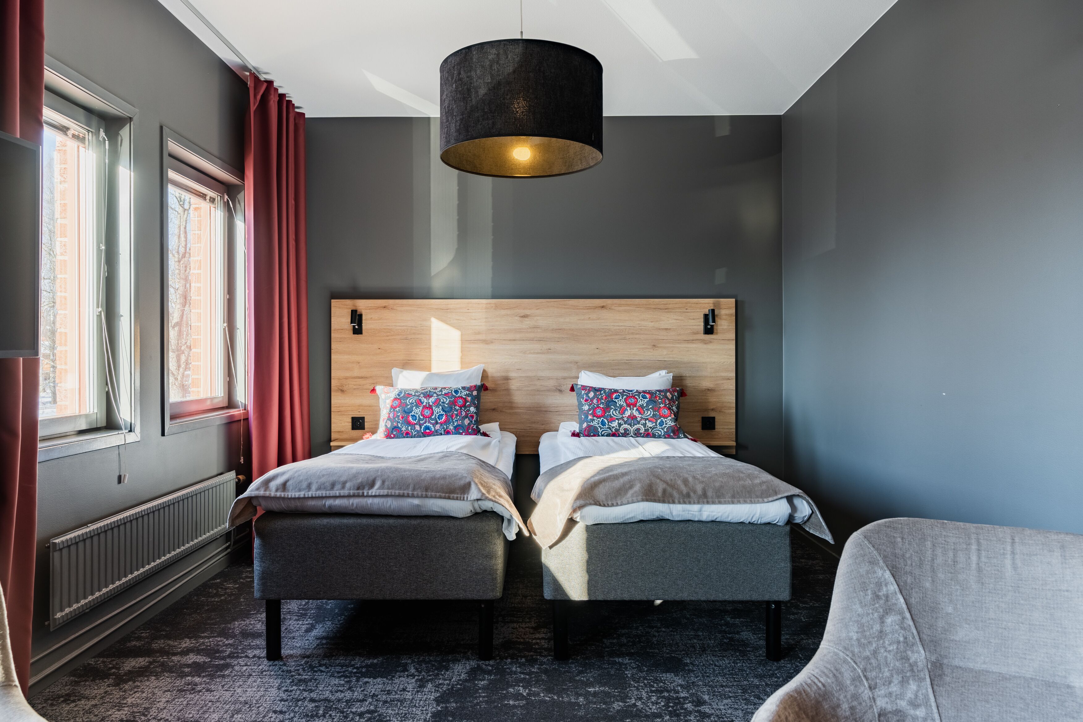 Ein modernes Hotelzimmer mit zwei Betten, einem Holz-Kopfteil, einem großen Fenster mit roten Vorhängen und einer schwarzen Pendelleuchte.