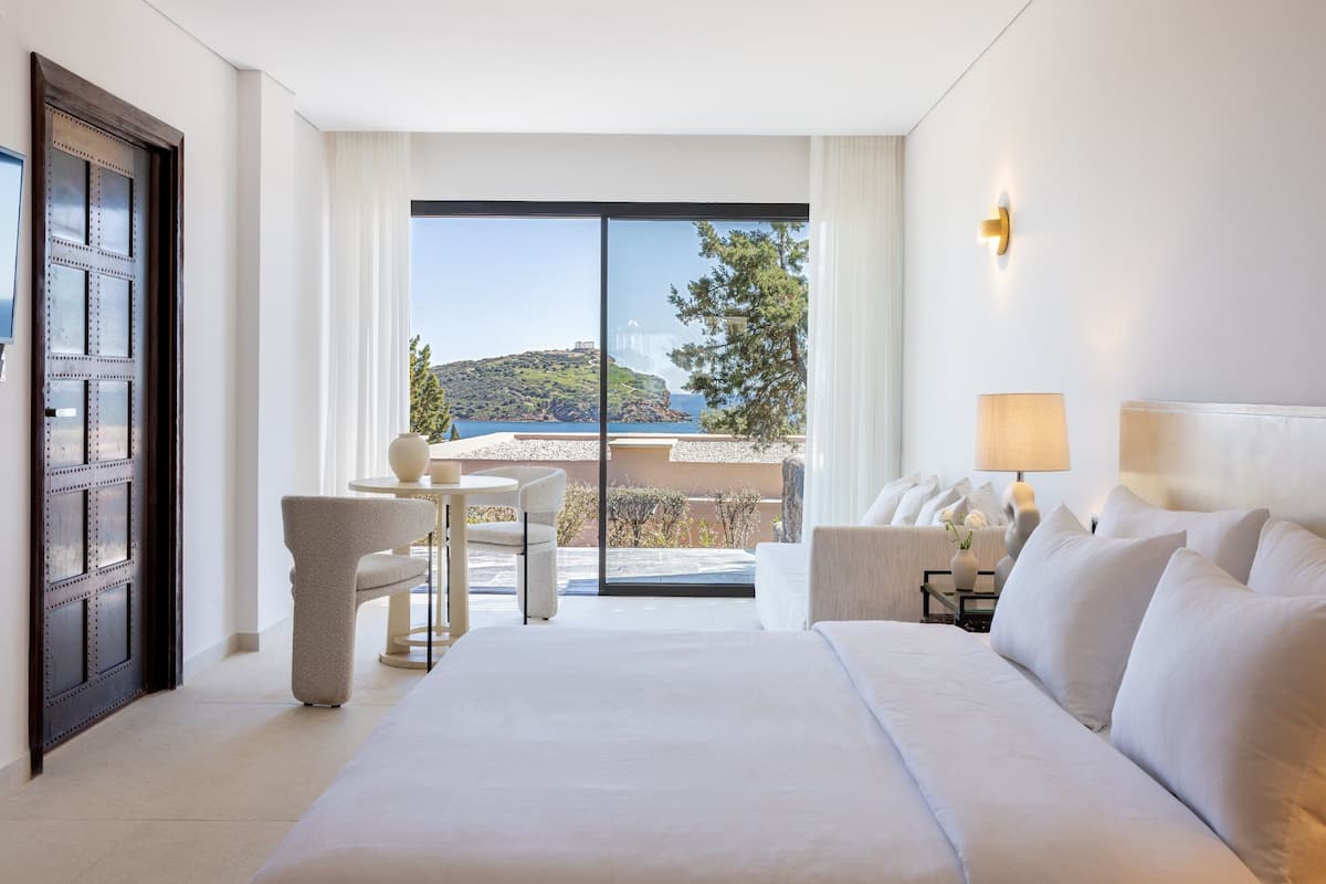 Ein Hotelzimmer mit einem großen Bett, einer Sitzecke mit Stühlen, einem Tisch und Blick auf das Meer und die Hügel.