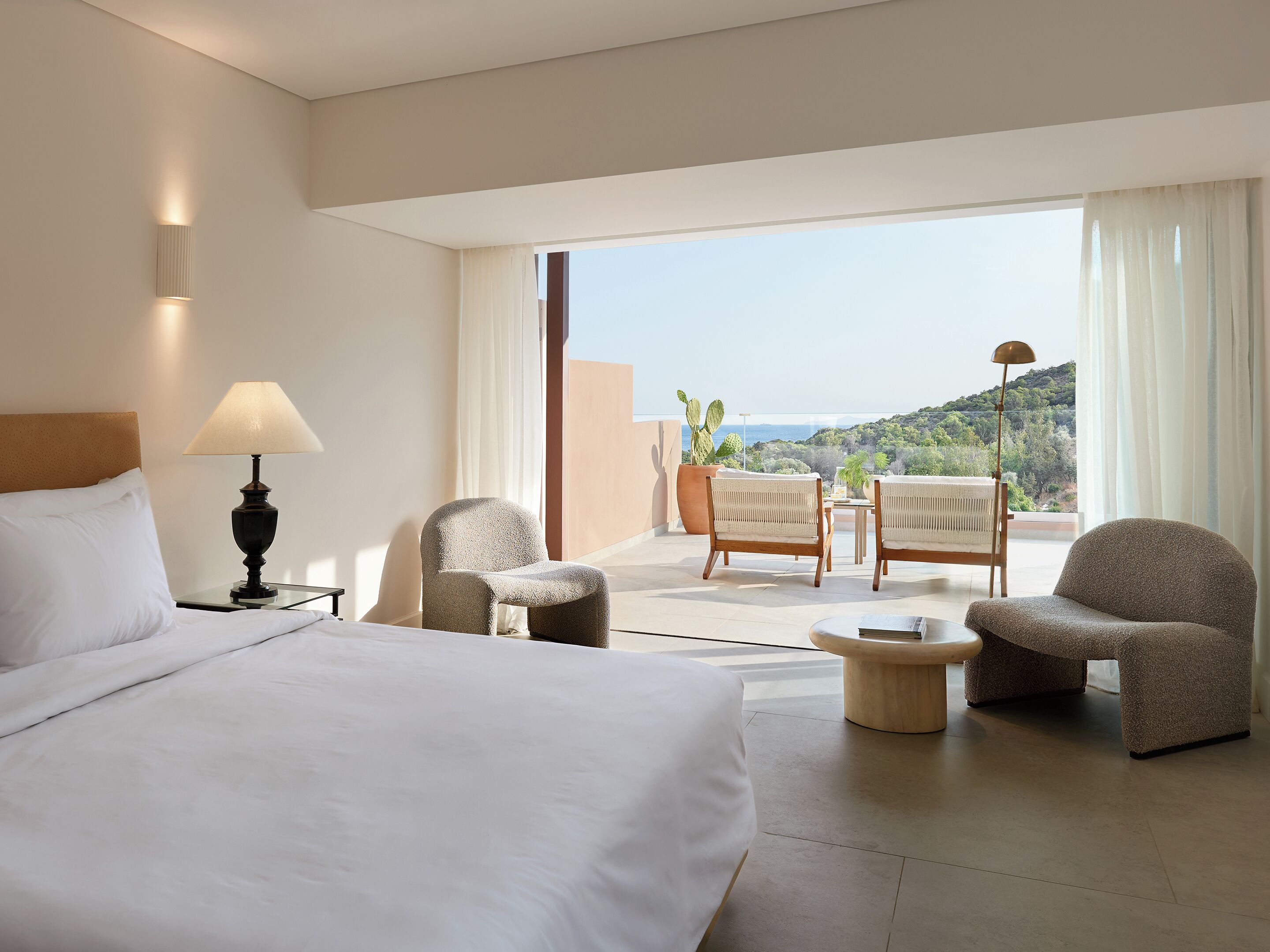 Une chambre d’hôtel avec un lit, une table de chevet avec une lampe, deux fauteuils, une petite table et une vue sur la mer par de grandes fenêtres.