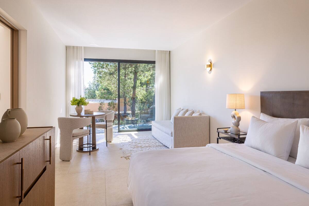 Ein modernes Hotelzimmer mit einem großen Bett, einer Essecke mit Tisch und Stühlen sowie Blick ins Freie durch eine Schiebetür.