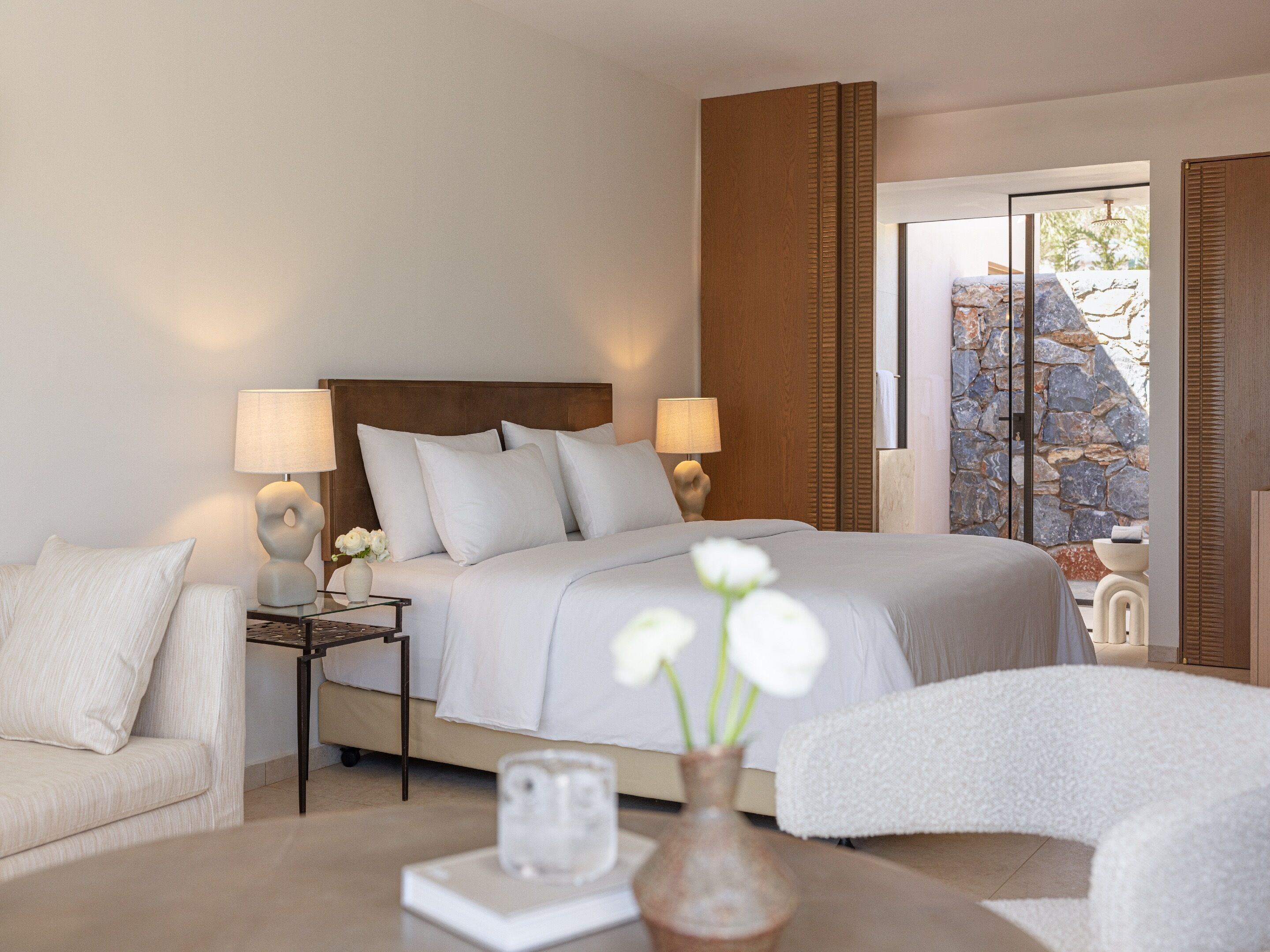 A modern bedroom with a large bed, two bedside lamps, a nightstand, a sofa, and a view of a stone wall through a glass door.
