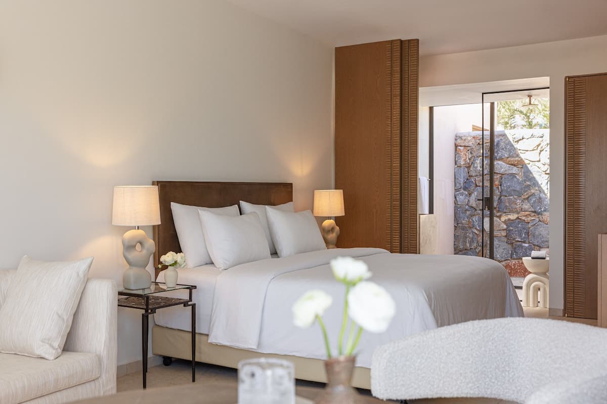 A modern bedroom with a large bed, two bedside lamps, a nightstand, a sofa, and a view of a stone wall through a glass door.