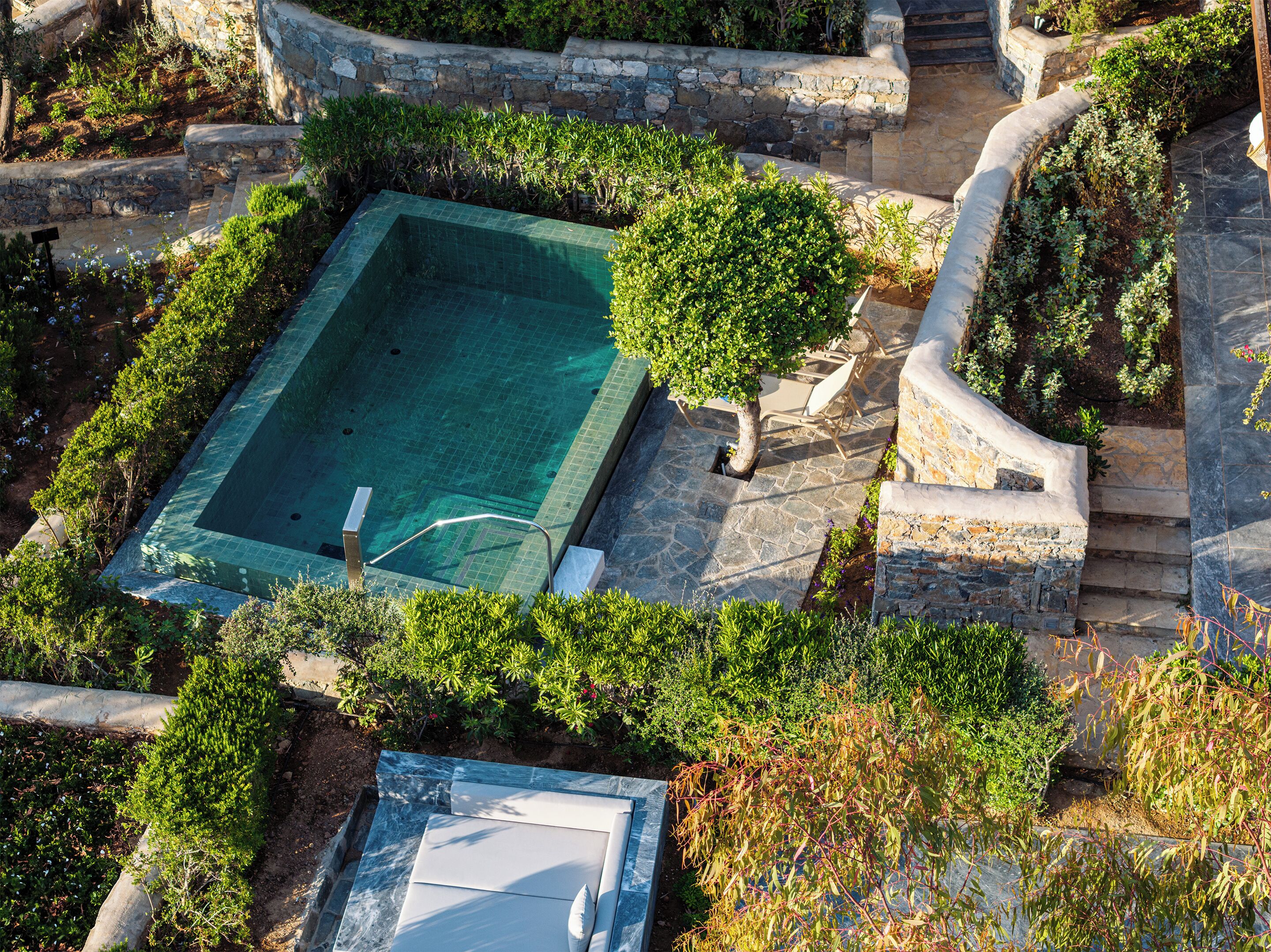 Una piscina rectangular rodeada de muros de piedra y vegetación, con escalones que llevan a ella.