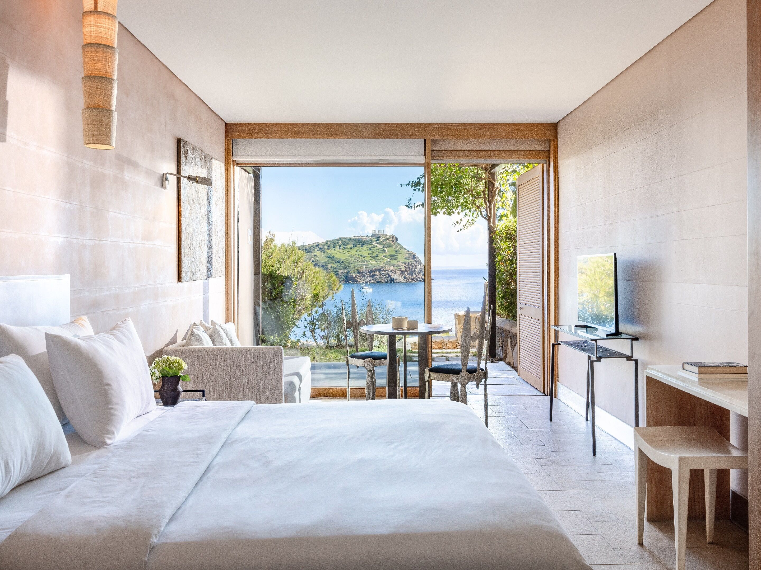 A modern hotel room with a large bed, a desk, a chair, and a television. There is a balcony with a view of the sea and a mountain.