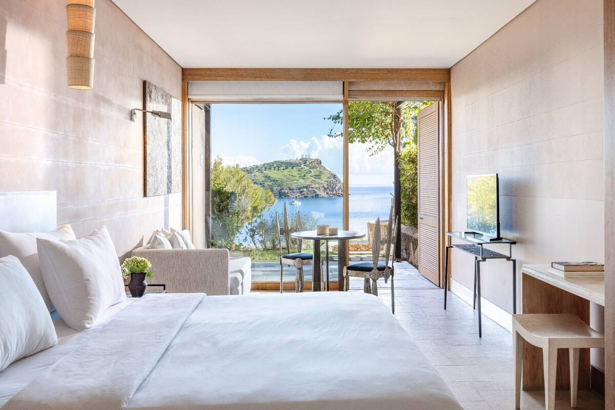 A modern hotel room with a large bed, a desk, a chair, and a television. There is a balcony with a view of the sea and a mountain.