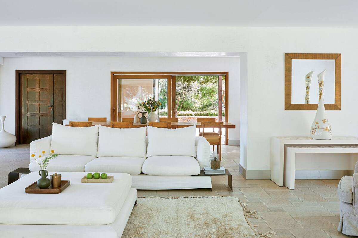 Ein modernes Wohnzimmer mit einem weißen Sofa, einem Holztisch und einem großen Fenster mit Blick auf Grünflächen.