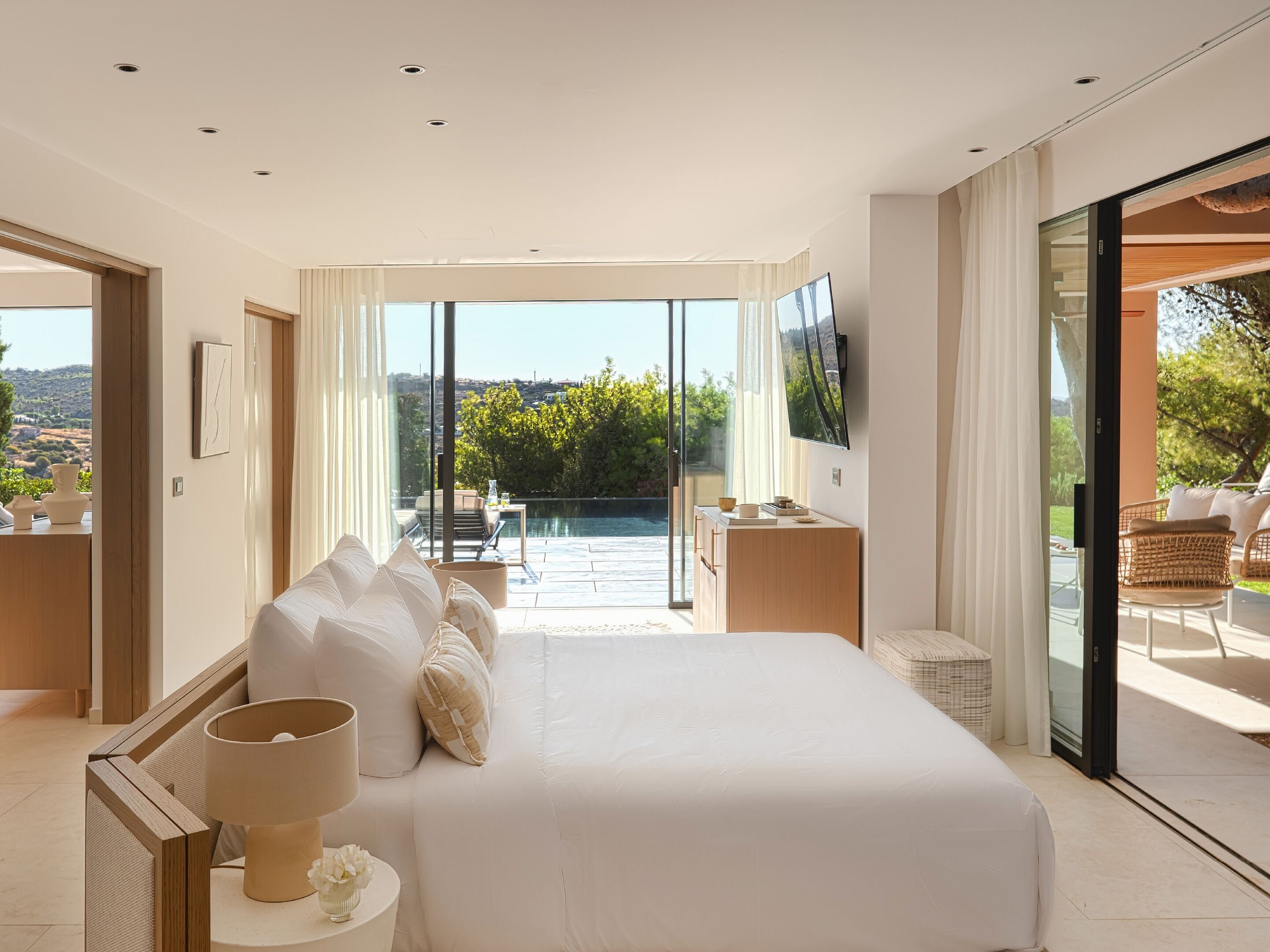 A modern bedroom with a large bed, a TV, and a balcony with a view of the landscape.