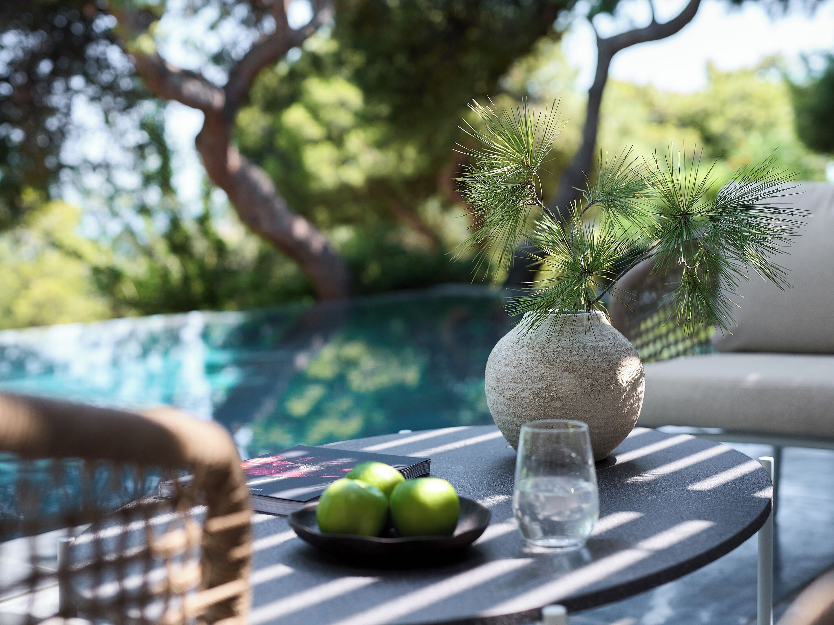 Una mesa redonda con un vaso de agua, un bol de manzanas verdes y una rama de pino decorativa en un florero texturizado.