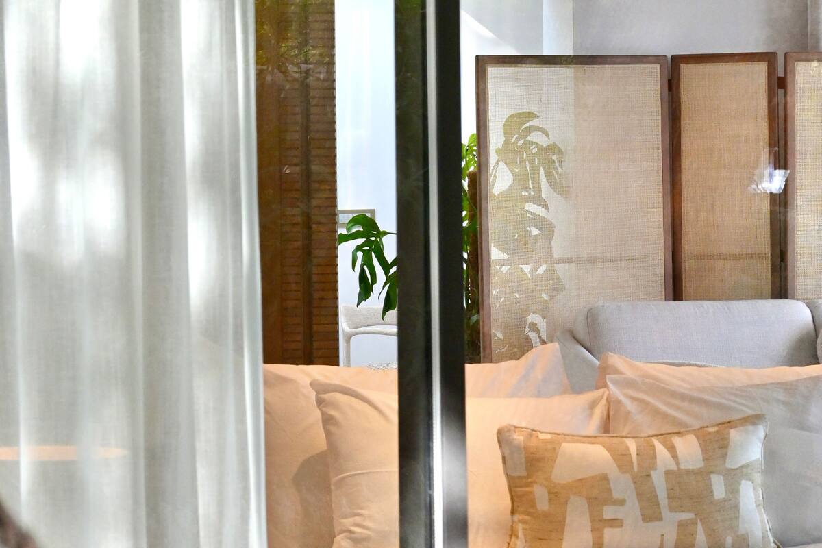 Ein modernes Wohnzimmer mit einer Couch, einem Kissen mit Muster und einem großen Fenster mit transparenten Vorhängen.