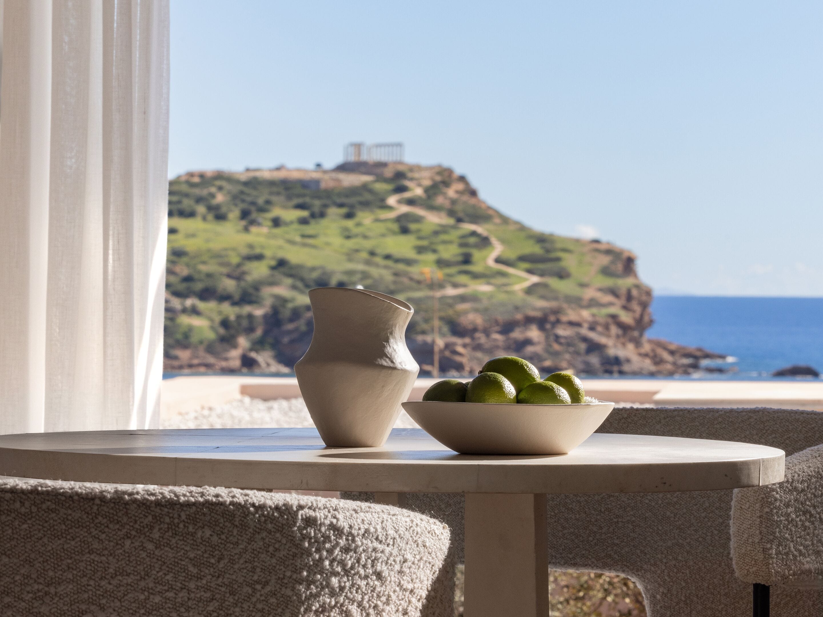Una mesa con un cuenco de limones y un florero, con vista a un paisaje costero con un acantilado y un faro.