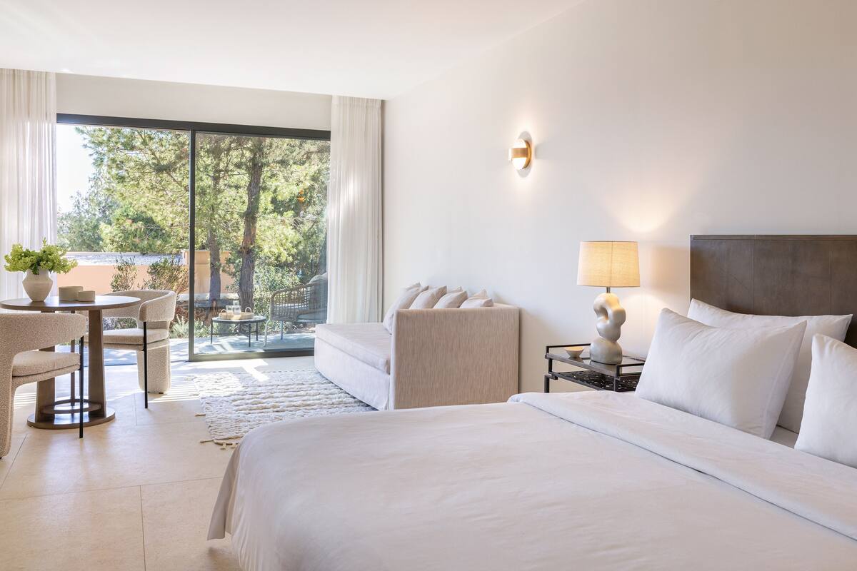 Ein Hotelzimmer mit einem großen Bett, einer Sitzecke mit Tisch und Stühlen, einer Lampe und Blick auf Bäume durch das Fenster.