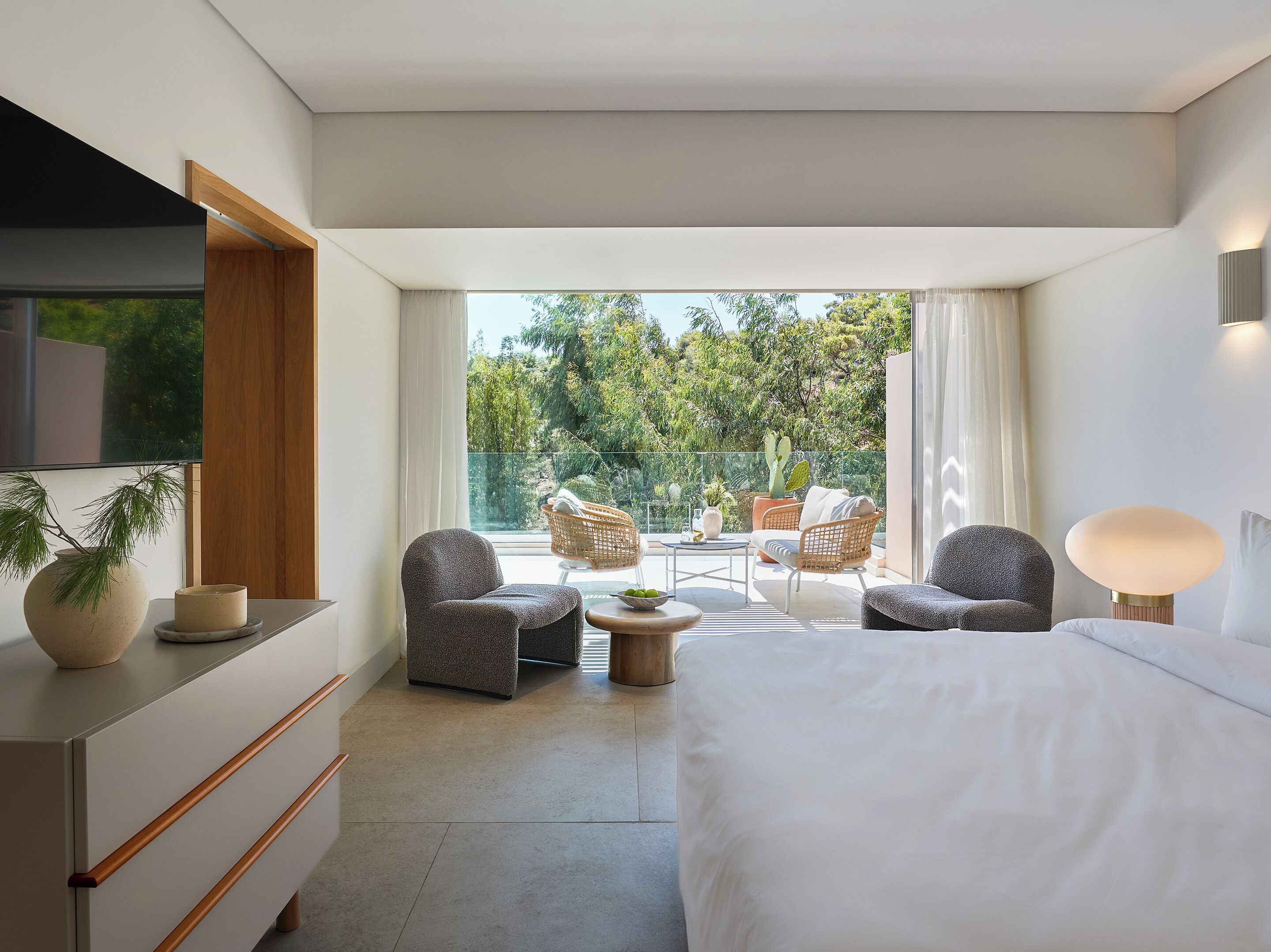 A modern hotel room with a large bed, two armchairs, a small table, and a TV. The room has a balcony with a view of trees and a fence.