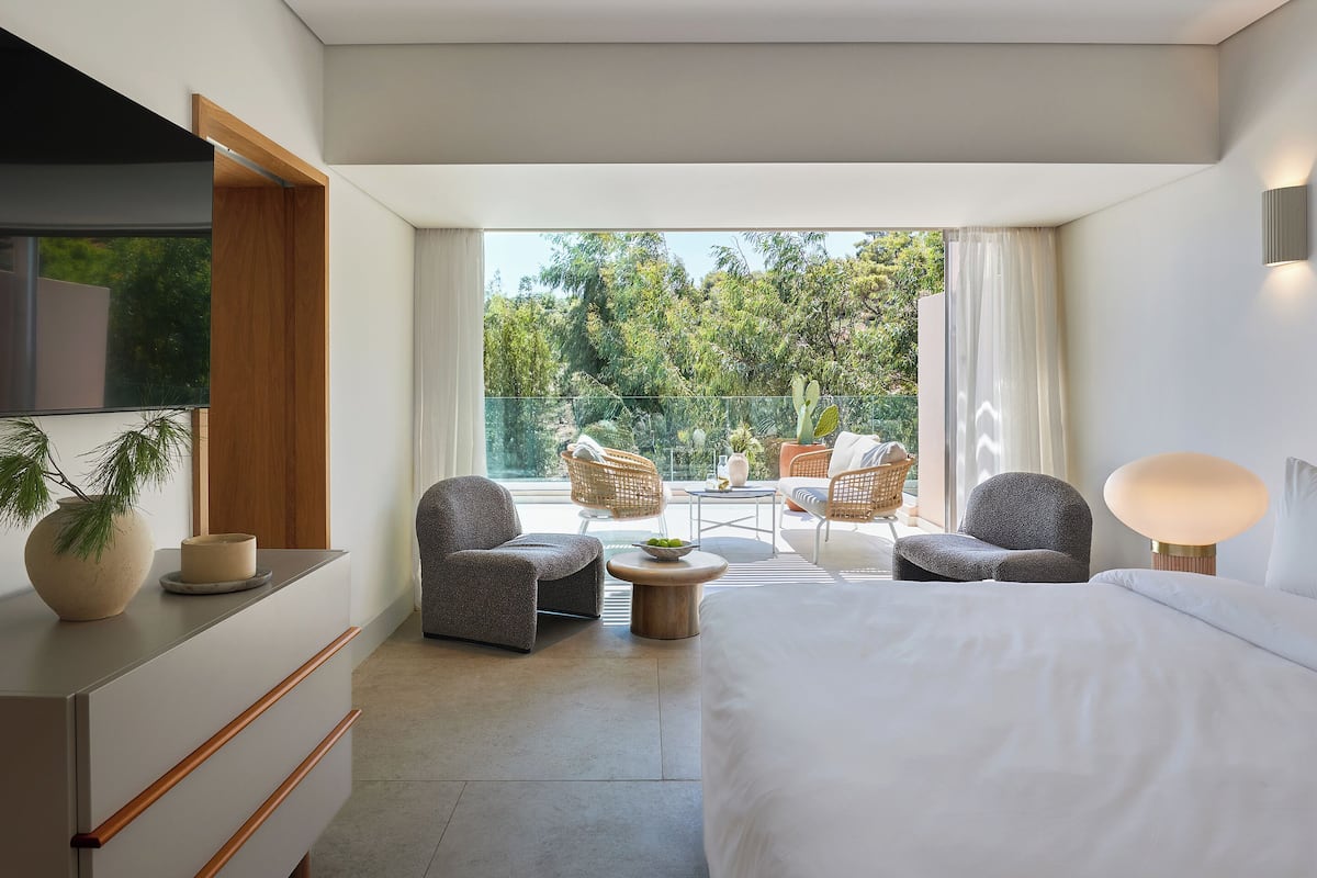 Une chambre d’hôtel moderne avec un grand lit, deux fauteuils, une petite table et un téléviseur. La chambre dispose d’un balcon avec vue sur les arbres et une clôture.