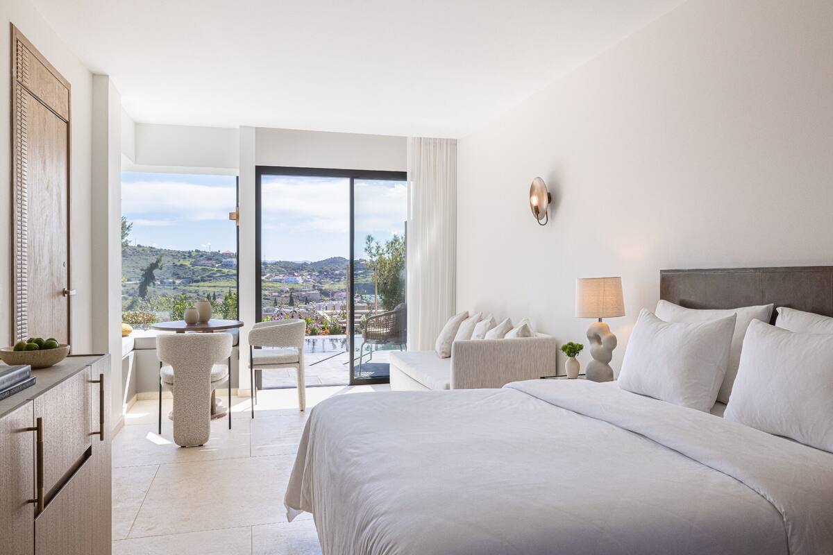 Ein modernes Hotelzimmer mit einem großen Bett, einem Balkon mit Blick auf die Stadt, einem Essbereich und Ausblick auf die Landschaft.