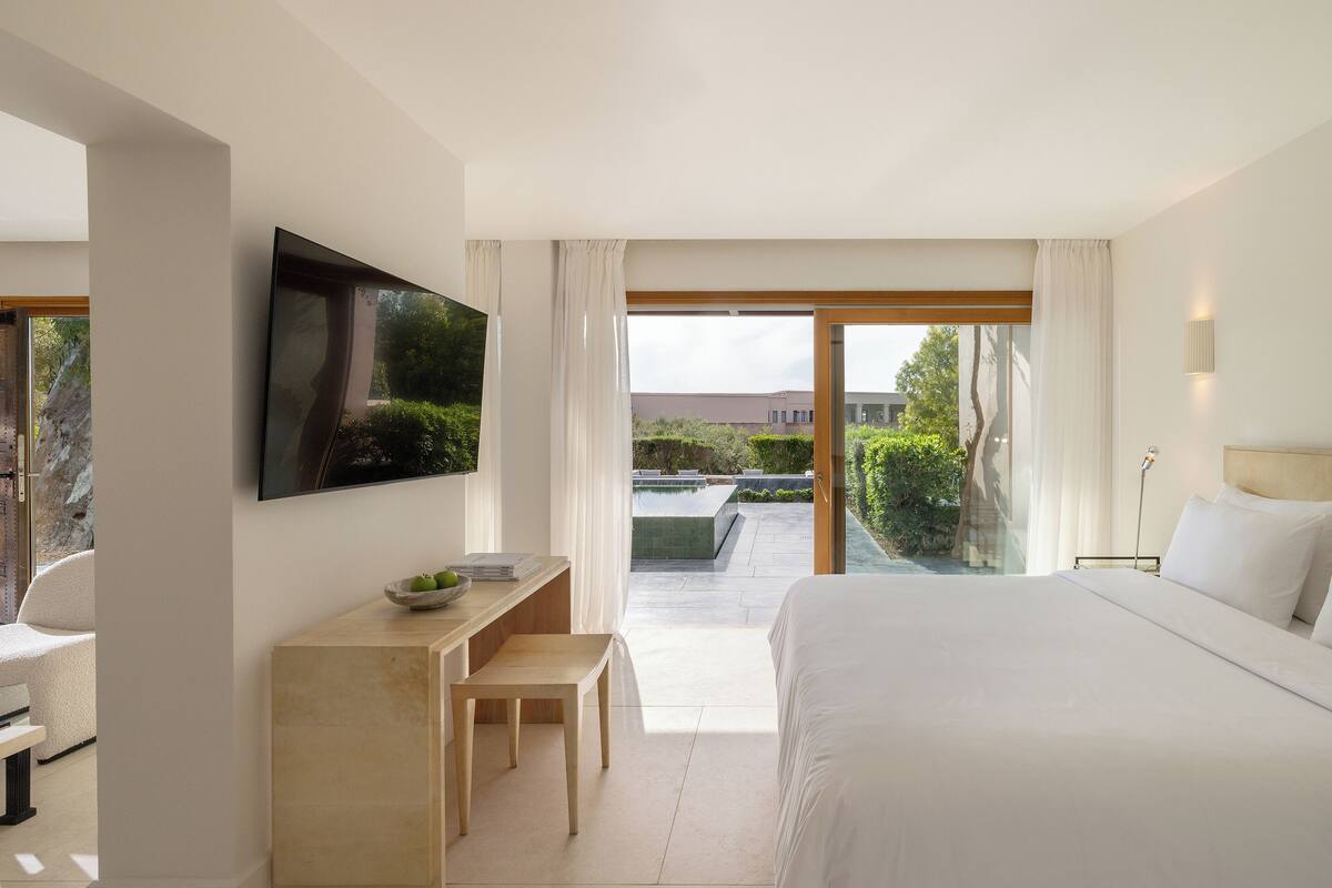 Ein modernes Schlafzimmer mit einem großen Bett, einem an der Wand montierten Fernseher, einem kleinen Tisch mit einer Obstschale und Blick auf einen Garten und Pool.