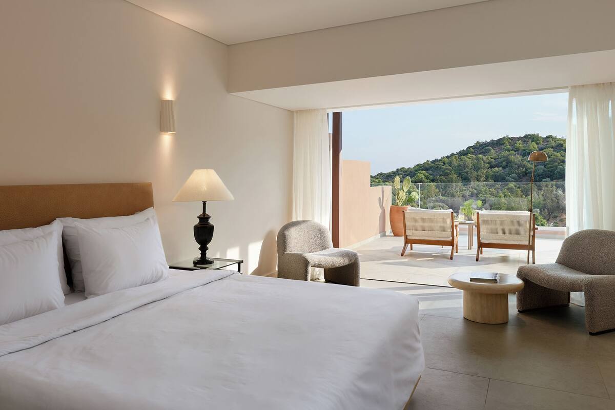Ein Hotelzimmer mit einem Bett, einem Nachttisch mit Lampe, zwei Sesseln, einem kleinen Tisch und einem Balkon mit Blick auf eine Hügel.'