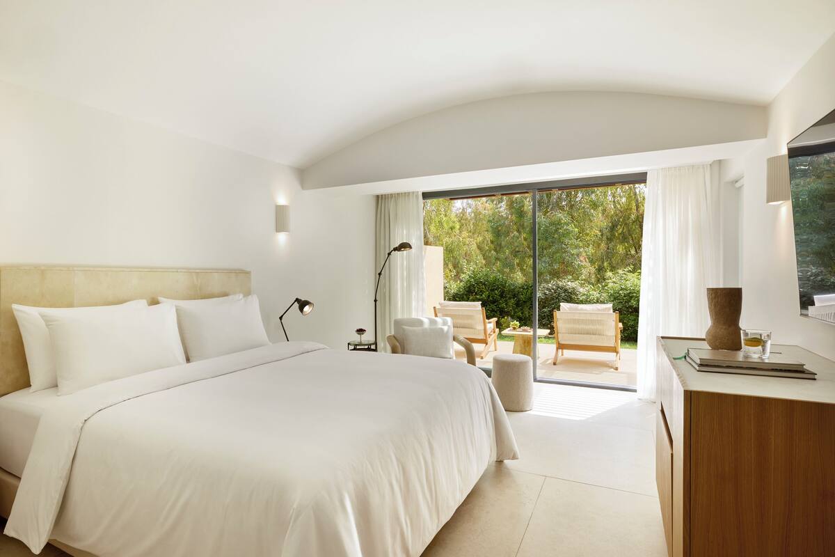 Ein modernes Hotelzimmer mit einem großen Bett, einem Schreibtisch und Blick auf einen Garten durch eine Schiebetür.