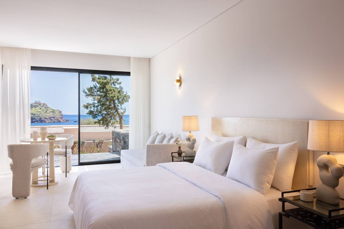 Ein Hotelzimmer mit einem großen Bett, zwei Nachttischen inklusive Lampen, Meerblick und einem Balkon mit Tisch und Stühlen.