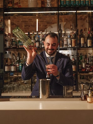 Un barman sostiene una botella y un vaso, con una barra repleta de diversas botellas al fondo.