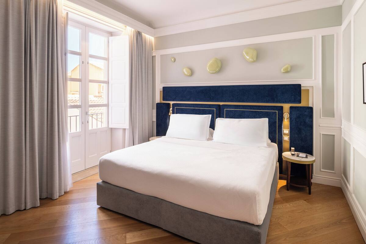Une chambre d’hôtel avec un grand lit, une table de chevet, une fenêtre avec des rideaux et un luminaire fixé au mur.