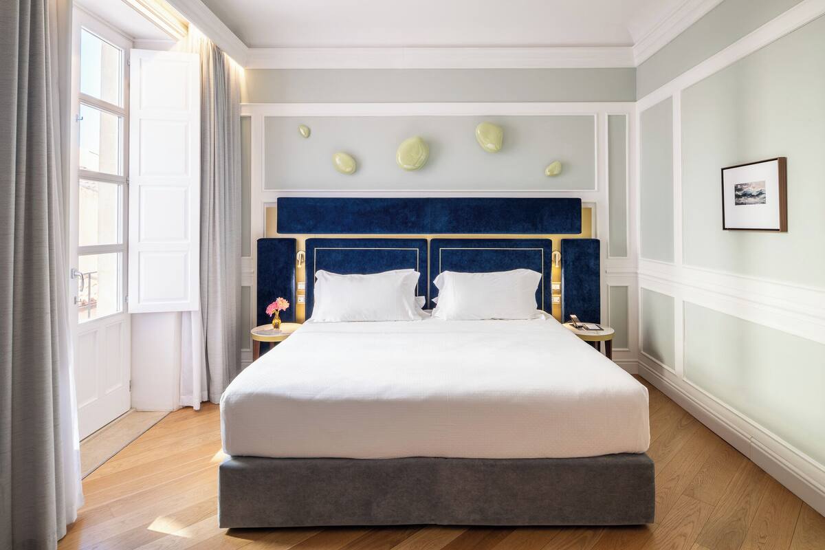 Une chambre d’hôtel avec un grand lit, deux tables de chevet, une fenêtre avec des rideaux et une œuvre d’art accrochée au mur.