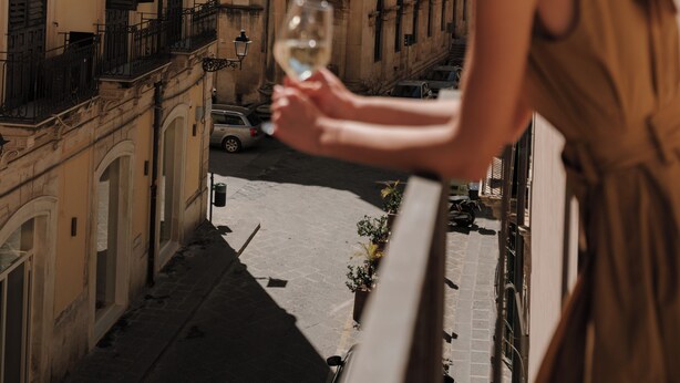 Una mujer sosteniendo una copa de vino en un balcón con vista a una calle con edificios históricos y una torre del reloj.