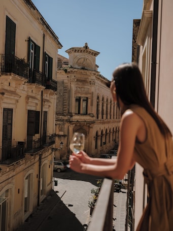 Una donna tiene un bicchiere di vino su un balcone con vista su una strada fiancheggiata da edifici storici e una torre dell'orologio.. Palazzo Artemide - VRetreats, SLH Collection