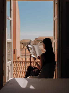 Una persona leyendo un libro en un balcón con vistas a edificios y un cielo despejado.