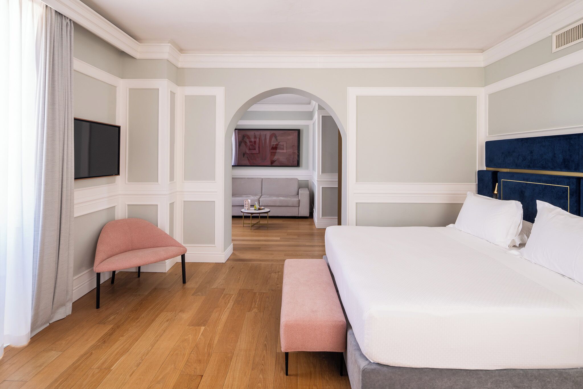 A hotel room with a bed, a bench, a TV, a sofa, and an arched doorway leading to another room.