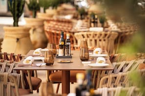 Mesa lista para comer con platos, vasos y botellas, rodeada de sillas de mimbre.