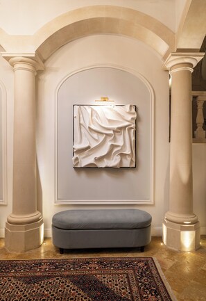 A room with a large, ornate column, a wall-mounted artwork, a grey ottoman, and a patterned rug. -  Palazzo Artemide - VRetreats, SLH Collection (Syracuse)