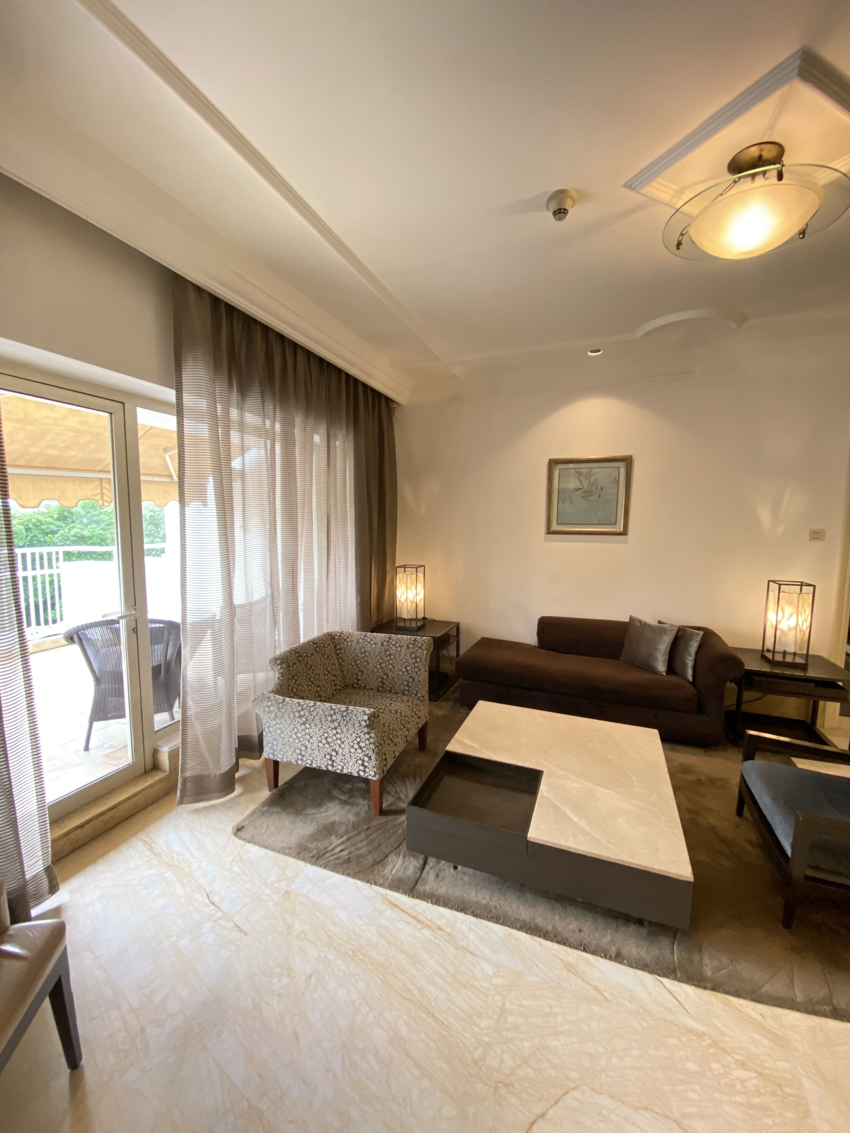 A modern living room with a sofa, armchair, and coffee table, a sliding glass door leading to an outdoor area, and a ceiling light fixture.