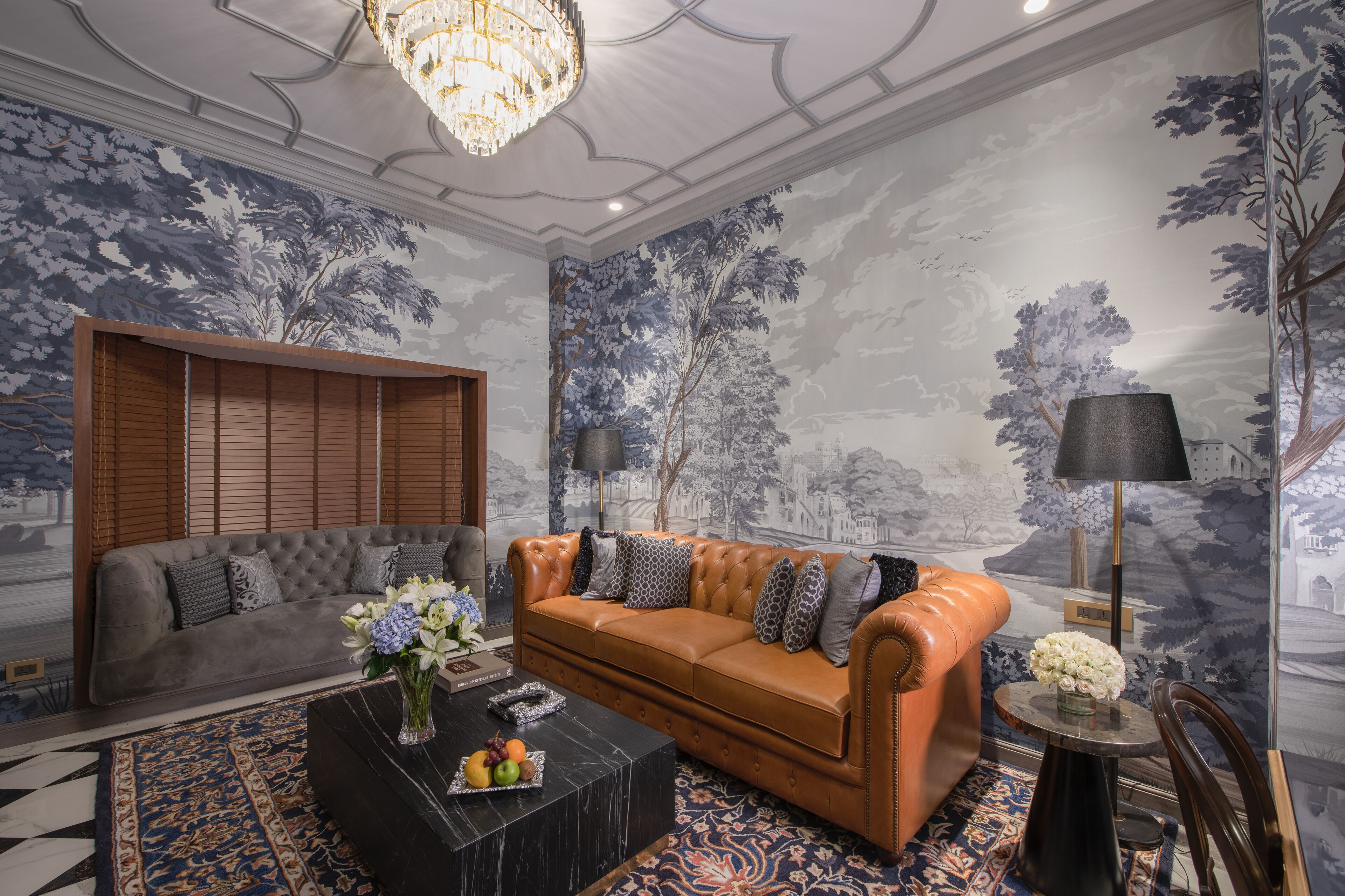 A living room with a brown leather sofa, a grey sofa, and a black coffee table with fruit and flowers. There are two floor lamps and a chandelier.